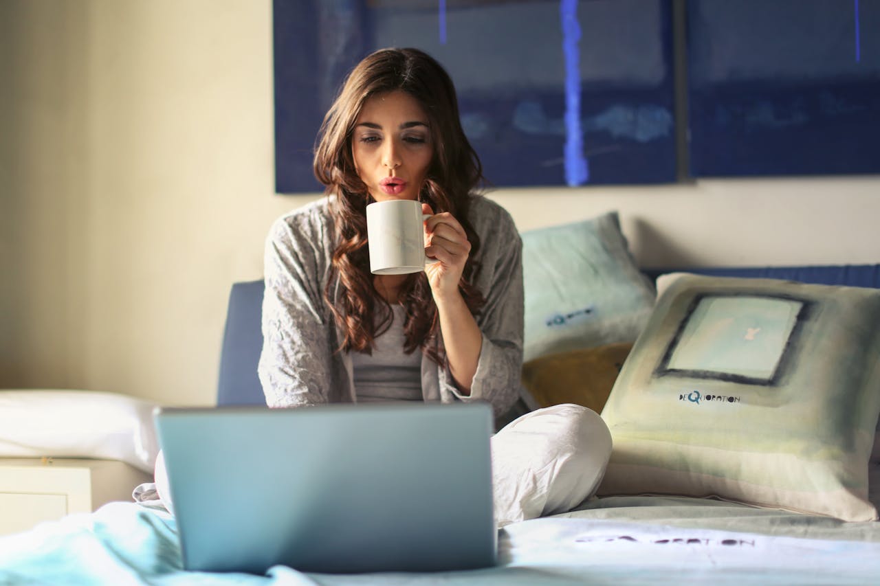 Person blowing on a cup of coffee, laptop on bed. Image by Pexels