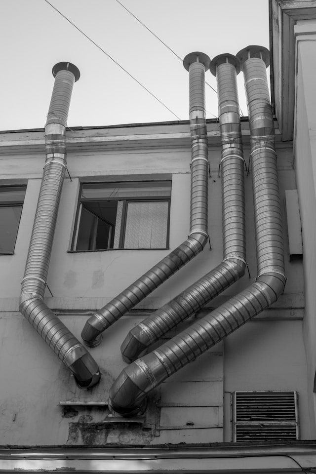A black and white photo showing multiple HVAC pipes and units on the side of a building. Image by Unsplash