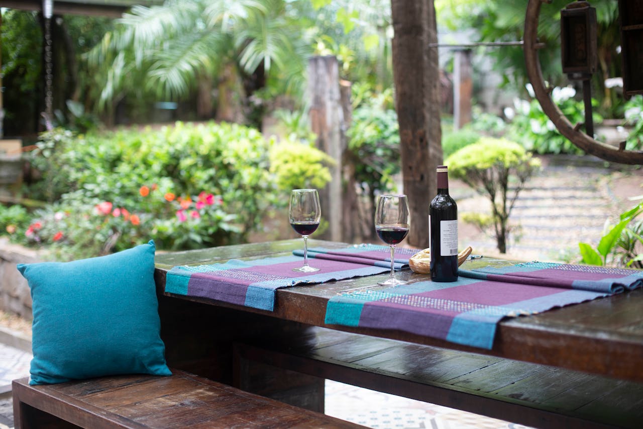 Wine bottle and two glasses of wine on a table outside. Lots of plants, trees and bushes. Image by Pexels