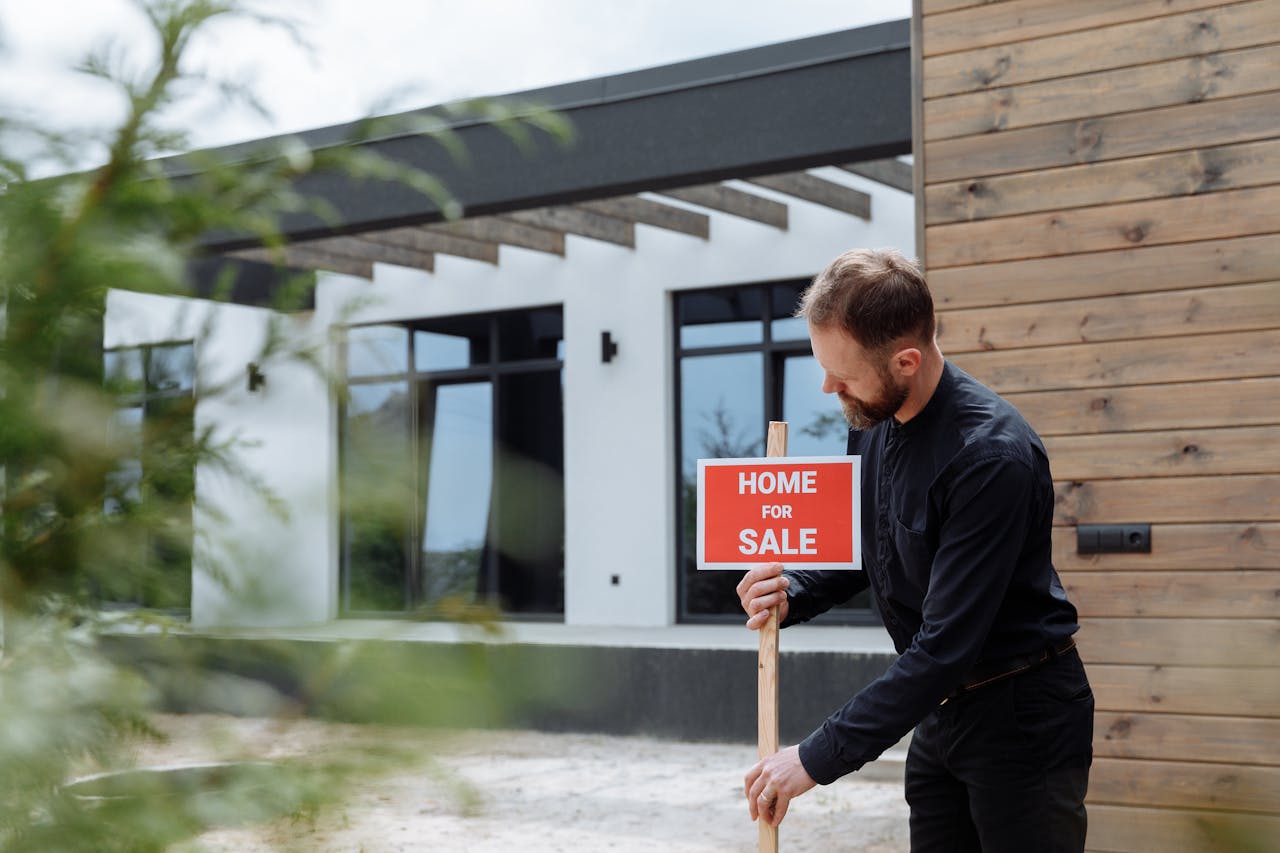 Person with a white hard hat putting a for sale sign in front of a white house. Image by Pexels.